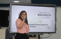 Foto de uma mulher branca, de cabelos claros, sorrindo e falando ao microfone que segura com a mão direita. Ela usa calça preta e blusa rosa claro, e está de pé, à frente de uma tela de projeção com fundo branco, onde se lê JANEIRO BRANCO e parte de outro texto, em tons de preto e cinza.