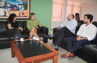 Foto de duas mulheres (à esquerda) e dois homens (à direita) sentados em sofás pretos, conversando. Vê-se, ao centro, uma mesa baixa de madeira e tampo preto com copos d'água e xícaras de café. Ao fundo, parede verde com quadro artístico colorido e persianas brancas.