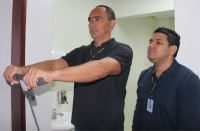 Foto de dois homens claros e de cabelos escuros, usando camisas escuras. À esquerda, um deles utiliza equipamento de exame de bioimpedância, enquanto o outro, à direita, observa os dados que estão sendo registrados no equipamento.