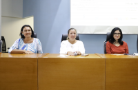  Mesa composta por três mulheres que estão posando para foto. A foto foi tirada dentro de um auditório com paredes azul e branca.