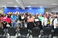 Foto de várias pessoas de pé, sorrindo e posando para a foto. À sua frente, veem-se cadeiras pretas de auditório. Ao fundo, parede azul.