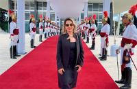 Foto de uma mulher branca, cabelos curtos e claros, usando óculos escuro, vestido e blaser preto, de pé sobre tapete vermelho na entrada do STF e ladeado por guardas usando o fardamento oficial dos Dragões da Independência.