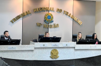 Foto da bancada principal de um plenário, com tampo de mármore branco e base de granito preto. Ao centro do tampo, vê-se o brasão da República do Brasil em dourado. Posicionadas na bancada, estão um homem claro à esquerda, uma mulher clara ao centro e outras duas mulheres claras à direita. Todos usam roupas pretas e olham para monitores de computador à sua frente. Ao fundo, parede listrada verticalmente em branco e cinza, contendo o brasão da República do Brasil e o nome do Tribunal  em dourado.