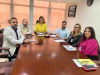 Seis pessoas posando para foto, três homens e três mulheres. Todos estão sentados apoiados em mesa de madeira com bloco de notas, papéis e canetas sobre a mesa.