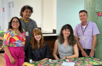 Foto de cinco pessoas, sendo uma mulher e dois homens em pé, e duas mulheres sentadas à uma mesa com toalha estampada. Todos sorriem, posando para a foto.