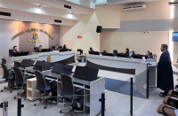 Foto panorâmica de um plenário com paredes em tons claros e teto com nichos e camadas de diferentes alturas. Veem-se as bancadas e cadeiras onde se posicional sete pessoas durante sessão de julgamento. À direita, vê-se um homem de pé ao microfone, usando toga preta.