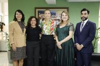 Cinco pessoas posando para foto, sendo quatro mulheres e um homem, dentro de uma sala. Ao fundo, parede verde com quadro colorido.