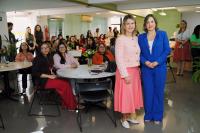 À frente, duas mulheres posando para foto, dentro de uma sala. Ao fundo, mulheres sentadas ao redor de mesas redondas e outras muheres em pé.