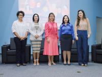 Foto de cinco mulheres de pé, lado a lado, posando e sorrindo para a câmera. Atrás delas, veem-se poltronas pretas e uma tela de projeção em uma parede azul, exibindo ilustração colorida.