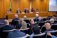 Foto de um auditório, com paredes ao fundo revestidas com madeira. Mesa solene composta por três pessoas, dois homens e uma mulher (ao centro). 