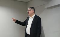 Homem branco, com cabelos grisalhos, vestido com terno preto e camisa branca, usando óculos de grau, dentro de uma sala com paredes brancas. Fazendo gesto explicativo com o braço esquerdo.