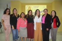 Foto de sete mulheres brancas, de pé, sorrindo para a câmera. Atrás delas, vê-se uma parede amarela onde está sendo projetada a arte do evento.