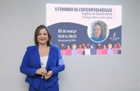 Mulher vestindo blaser e calça na cor azul, posando para foto. Ao fundo, imagem projetada de um datashow com detalhes da palestra ministrada.