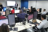 Foto tirada dentro de uma sala, com paredes brancas e azuis. Ao fundo, um homem fazendo uma apresentação em frente à uma tela de led e pessoas sentadas em cadeiras, usando computadores que estão em cima de mesas.  