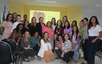 Foto de um grupo de pessoas sorrindo para a foto, parte delas agachada à frente das demais. Atrás de todas elas, vê-se uma parede amarela onde está sendo projetada uma arte do evento.