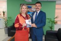 Duas pessoas posando para a foto, sendo uma mulher e um homem, dentro de uma sala, com parede de fundo esverdeada. Amulher usa saiade cor laranja e camisa social em tons terrosos. O homem usa terno e calça social em tom de azul. Eles seguram, ao centro, a Carta de Serviços da AGU-MA.