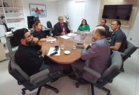 Sete pessoas sentados em torno de uma mesa redonda, dentro de uma sala. Ao fundo, uma televisão.