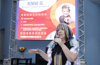 Foto de mulher branca, falando ao microfone, que segura com a mão esquerda e gesticula com a direita. Ao fundo, vê-se tela de projeção com arte da palestra.