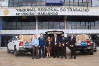 Foto de seis pessoas de pé e sorrindo para a câmera. Elas estão à frente de três caminhonetes carregadas de doações. Ao fundo, fachada do prédio-sede do TRT-16.