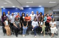Grupo de pessoas posando para uma foto dentro de um auditório. Parte das pessoas estão em pé, lado a lado, e outra parte estão sentadas em cadeiras de cor preta.