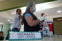 uma mulher, usando luvas com uma agulha na mão. À frente, placa escrita VACINAÇÃO INFLUENZA.