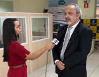 Duas pessoas em pé, uma mulher à esquerda e um homem. A mulher está entrevistando-o dentro de uma sala.