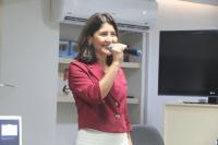 Uma mulher, vestida com camisa vinho e calça branca, segurando com a mão direita o microfone.