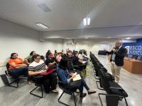 Foto tirada de um auditório com paredes brancas, contendo pessoas sentadas, prestando atenção ao que um homem fala à frente.