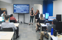 Foto panorâmica de uma sala de aula, onde os alunos são vistos de costas e há duas mulheres de pé, ao lado de uma tela de projeção. Ao fundo, parede branca com detalhe de faixa azul na vertical.