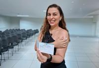 mulher posando para foto dentro de uma sala, segurando ciom a mão esquerda o cartão de vacina e com a mão direita apoiando o algodão no braço que foi aplicado a vacina.