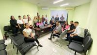 Grupo de pessoas posando para foto dentro de uma sala. O ambiente possui paredes de cor verde, cadeiras pretas e mesa de madeira.
