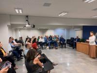  Imagem de um auditório com pessoas sentadas em cadeiras e ao centro uma mulher.