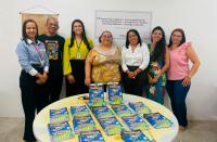 Sete pessoas em pé, atrás de uma mesa redonda, posando para foto. Sobre a mesa, cartilhas espalhadas.