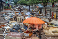 Imagem de uma pessoa, abaixada, segurando um guarda chuva, na calçada, no meio dos destroços causados pela enchente no RS. 