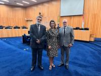 Trêsq pessoas posando para foto dentro de um plenário. Uma mulher ao centro, ladeada por dois homens.
