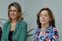 Foto de duas mulheres claras à frente de uma parede branca. A da esquerda está sorrindo e usa óculos e blazer verde. A da direita tem semblante sereno e usa vestido com estampa geométrica colorida.