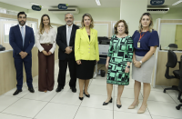 Foto de seis pessoas, sendo dois homens e quatro mulheres, de pé, sorrindo e posando para a câmera. Elas estão em um ambiente claro com divisórias nas laterais e placas de identificação próximas ao teto.