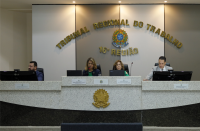 Foto de dois homens e duas mulheres sentados em uma bancada com computadores e microfones à sua frente. Ao fundo, parede listrada em branco e marrom, com o nome do TRT-16 em letras douradas, tendo abaixo o brasão da República. Na base da foto, vê-se a frente da bancada em mármore preto e branco com o brasão da República centralizado, todo em dourado.