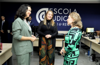Foto de três mulheres conversando e sorrindo. Atrás delas, veem-se mesas com computadores e, ao fundo, parede azul escura com a logomarca da Escola Judicial do TRT-16.