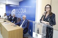 Foto de uma mulher em pé, atrás de um púlpito transparente, à direita, usando óculos e vestido escuro, e falando ao microfone, que segura com a mão direita. À esquerda, 4 pessoas sentadas a uma mesa de madeira, tendo às suas costas parede azul com a inscrição ESCOLA JUDICIAL em letras brancas.