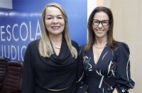 Foto de duas mulheres, uma loura e outra morena. Ambas usam roupa escura e posam para a câmera, sorrindo. Atrás delas, parede azul, onde se lê ESCOLA JUDICIAL em letras brancas.