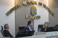 Foto de dois homens e uma mulher sentados numa bancada de tribunal, onde a mulher fala ao microfone, olhando para os homens e estes estão atentos à sua fala. Atrás deles, parede em tons de branco e bege, com letras douradas e o brasão da República.