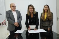 Foto com a presidente Márcia Andrea Farias assinando o edital de acordo direto. Ao lado da presidente estão o procurador Geral do Estado Valdêncio Caminha e a juíza Joanna D´Arck. 