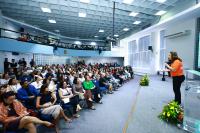 Foto mostra pessoas sentadas nas cadeira de um auditório, acompanhando uma palestra. Ao palco, uma mulher ministra a palestra. 