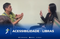Foto mostra, à esquerda, um homem claro de cabelos escuros, usando camisa verde e se comunicando por meio de sinais com as mãos. À direita, vê-se mulher clara de cabelos escuros, compridos e lisos, usando blusa e blazer pretos. Ela conversa em LIBRAS com o homem. Ambos estão sentados em frente a uma parede bege. Abaixo, tarja azul degradê, onde se vê a logomarca do TRT-16 e o texto branco ACESSIBILIDADE - LIBRAS. Abaixo, estão os endereços do TRT-16 nas redes sociais.