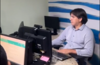 Foto mostra homem claro de cabelos escuros e lisos, usando camisa azul claro e utilizando um computador. Ele está sentado à frente de uma parede listrada de azul e branco.