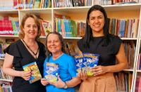 Foto mostra, três mulheres segurando as cartilhas educativas do programa TRT na Escolha. Ao fundo imagens de livros colocados em prateleiras 