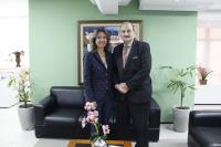 Foto mostra uma mulher e um homem. Ambos estão de pé. Ao fundo um ambiente de gabinete com sofá, quadros na paredes.