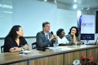 A imagem mostra quatro pessoas sentadas a uma mesa. Da esquerda para a direita, estão: uma mulher de cabelo preto; um homem de terno, usando um microfone; uma mulher negra, com cabelo curto e blusa clara; e uma mulher com cabelo preto. A mesa tem copos de água, microfones e papéis