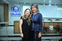 Foto mostra duas mulheres claras, de pé, lado a lado, sorrindo. Atrás delas ambiente de auditório.